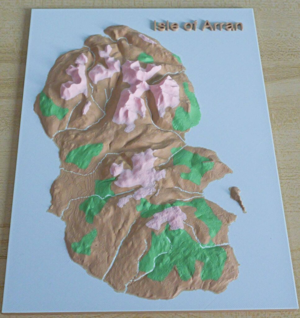 A 3D printed relief map of the Isle of Arran viewed from the south, showing the island's full terrain at true scale. The sea is shown in blue, with layered colour bands rising through green and brown to mark the island's peaks, including Goatfell. The map is printed in PLA and hand-finished.
